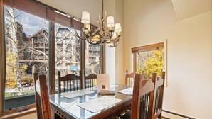 Comedor con mesa de vidrio, sillas de madera, una araña y grandes ventanales que ofrecen vistas a edificios y árboles.
