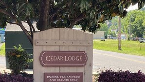 Een houten bord met de tekst 'CEDAR LODGE' en 'ALLEEN PARKEOREN VOOR EIGENAARS EN GASTEN', omringd door planten en een boom.