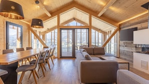 Une pièce spacieuse avec un plafond haut, des poutres apparentes, une longue table à manger entourée de chaises, un canapé et un téléviseur fixé au mur.