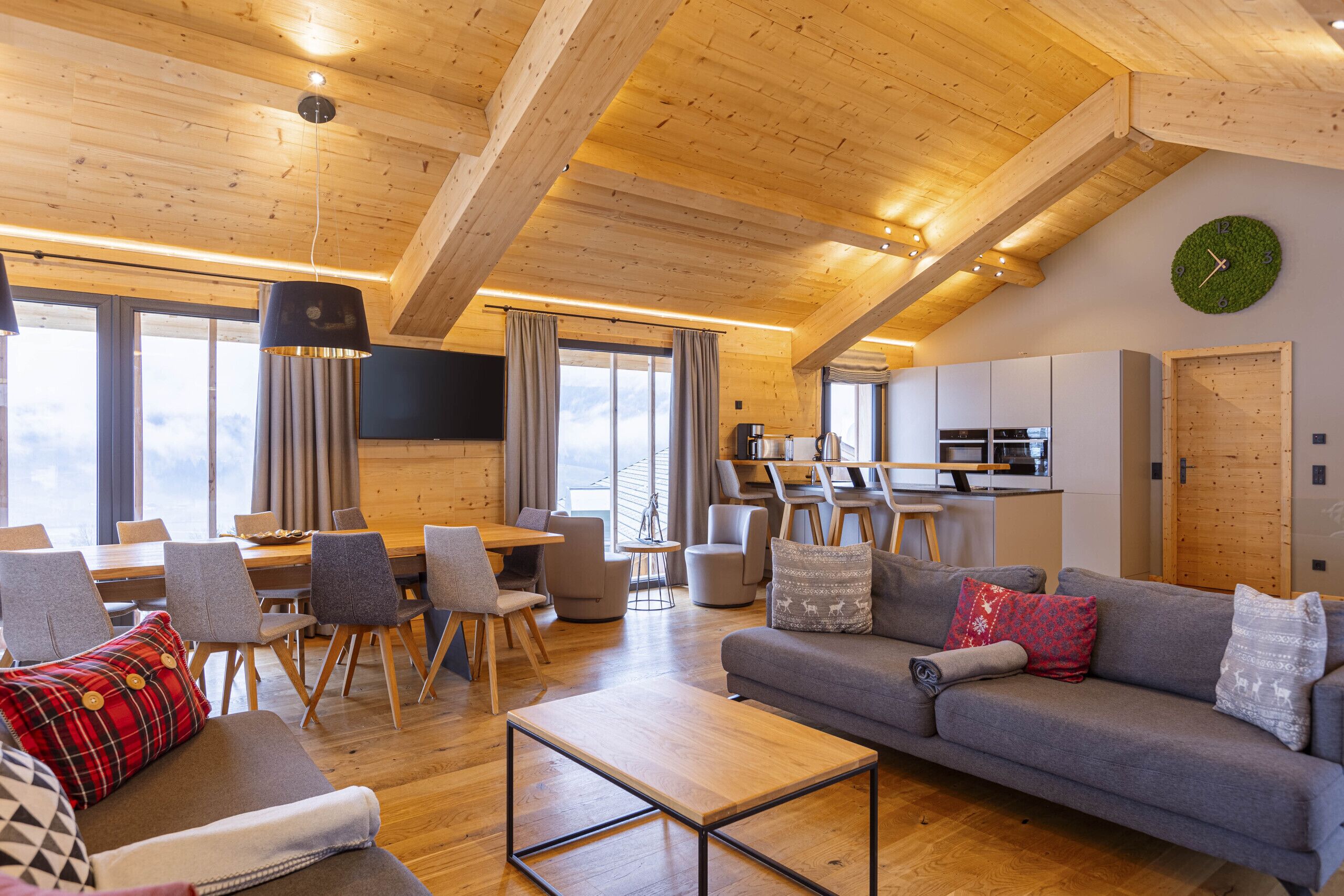Un vaste espace de vie doté d’un plafond en bois, d’un canapé gris, d’une table à manger avec des chaises, et d’une cuisine à l’arrière-plan.