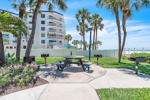 A picnic table with benches, surrounded by palm trees and a building with balconies. - Sand Dollar 107 Gorgeous Beachfront Views, Spacious, Pool, BBQ, Private Pier (Indian Shores)
