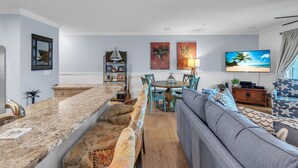 A modern living room with a kitchen area, a dining table, and a large flat-screen TV. - Oceanside302 Beachfront- Gorgeous Gulf Views, lots of updates, great location (Indian Rocks Beach)