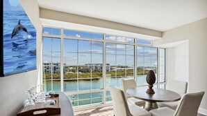 A dining area with a large window overlooking a body of water and buildings. There is a table with chairs and a tray of glasses and a remote control on the floor.