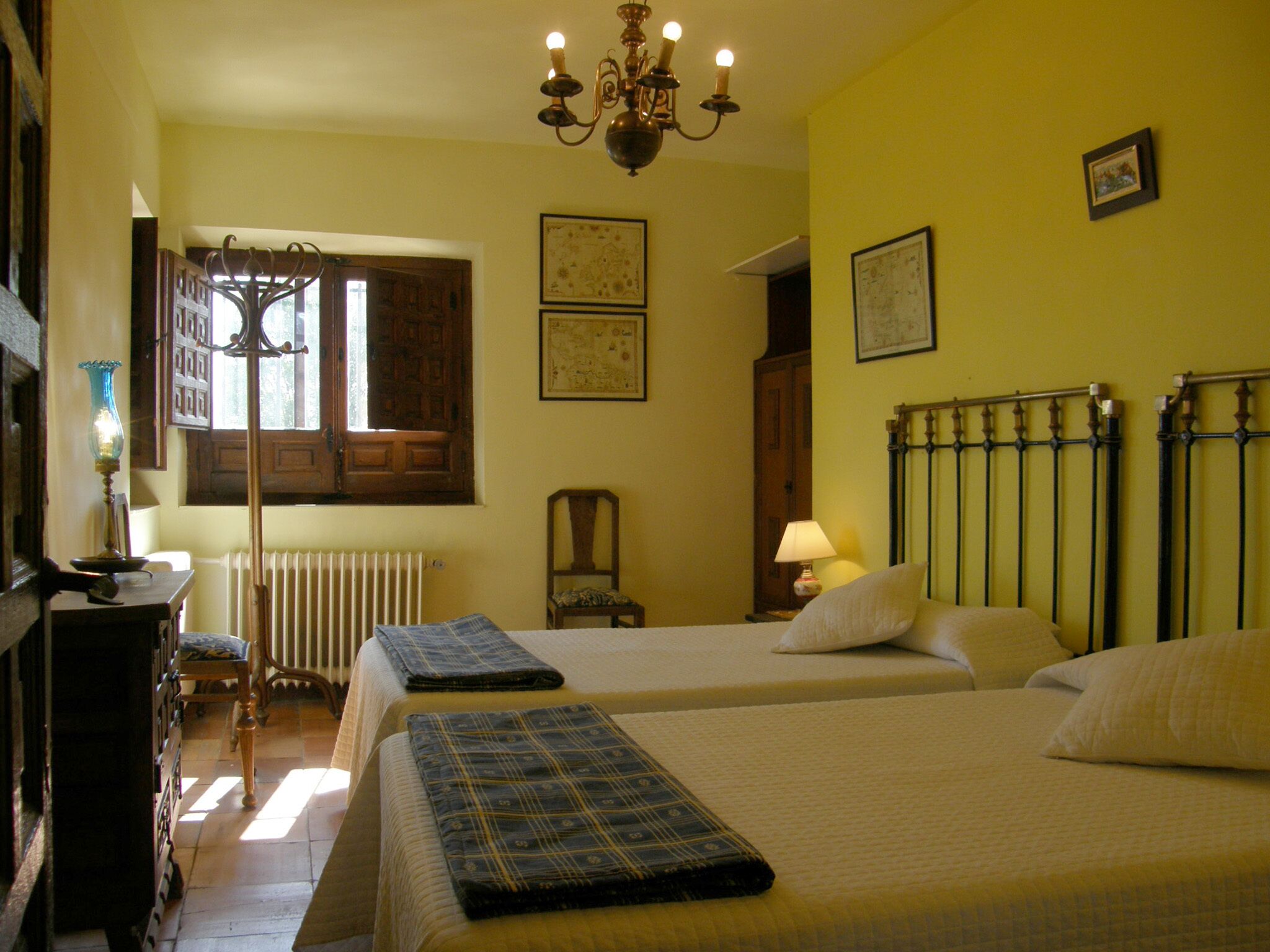 Quarto de hotel com duas camas, um radiador, uma cadeira de madeira, uma escrivaninha com uma lâmpada e um candelabro.
