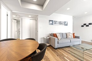 A modern living room with a round wooden dining table, a grey sofa with patterned cushions, and a glass coffee table.