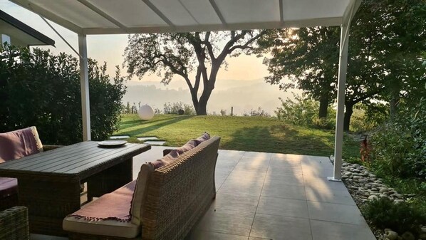A covered patio with a view of a lake, a wicker sofa, and a dining table.