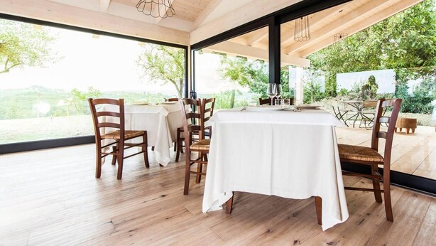 A dining area with wooden tables and chairs, large windows, and a view of greenery.
