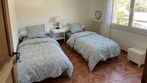 Une chambre avec deux lits, une table de chevet avec une lampe, un radiateur et une fenêtre avec des stores.