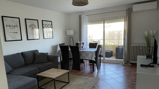 Un salon moderne avec un canapé, une table basse et un coin repas comprenant des chaises et une table. Des tableaux encadrés ornent le mur, et une fenêtre est agrémentée de rideaux.