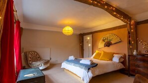 Ein Schlafzimmer mit einem Bett, einem Sessel, einer hölzernen Kommode, einem Ventilator und einem Koffer.
