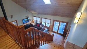 A spacious loft with a wooden staircase, a living area with a sofa and TV, large windows, and a wooden ceiling. - El Cabitan: 4BR Scenic Mountain Views, Hot Tub, Game Room & Fire Pit (West Jefferson)