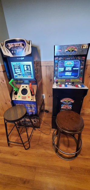 Deux bornes d’arcade, un tabouret et une table dans une pièce dont le sol est recouvert de planches de bois.
