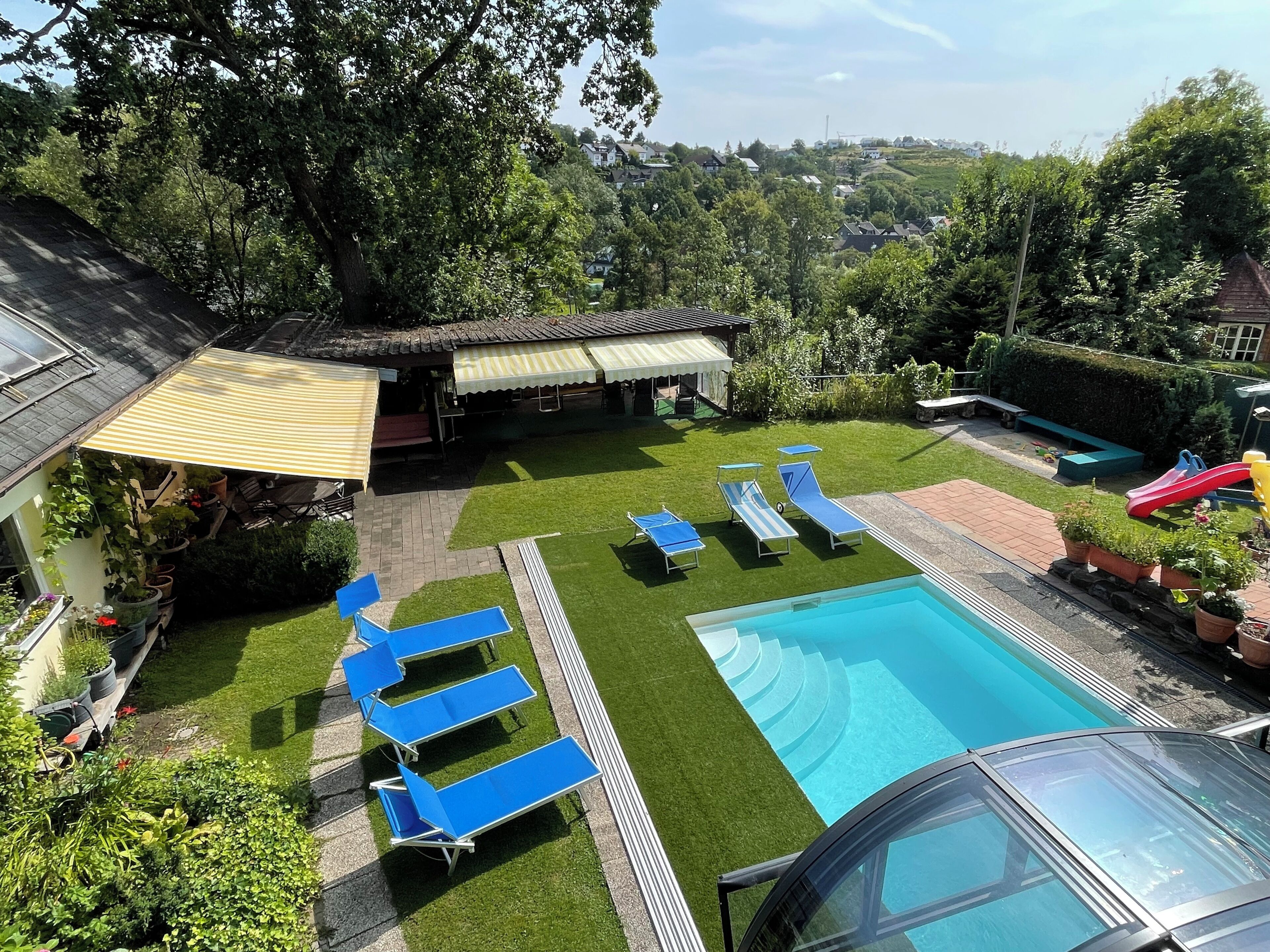 A backyard with a swimming pool, sun loungers, a slide, and a covered patio area.