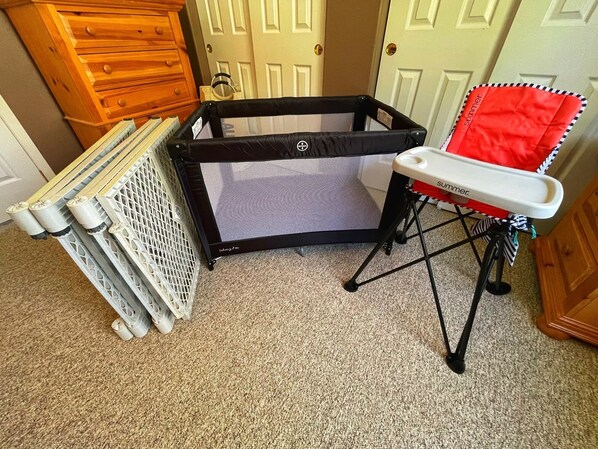 A portable crib with a mesh screen, a red and white high chair, and a wooden dresser in a room with carpeted flooring.