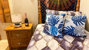 Ein ordentlich bezogenes Bett mit einem gemusterten Überwurf, ein Nachttisch mit einer Weinflasche, einer Oliven-Schale und einer Lampe.