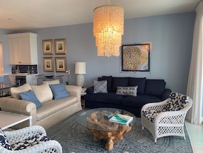 Uma sala de estar com um sofá bege, um sofá azul escuro, uma mesa de centro de vidro e uma poltrona de vime branca. Há um candelabro pendurado no teto e uma obra de arte emoldurada na parede.