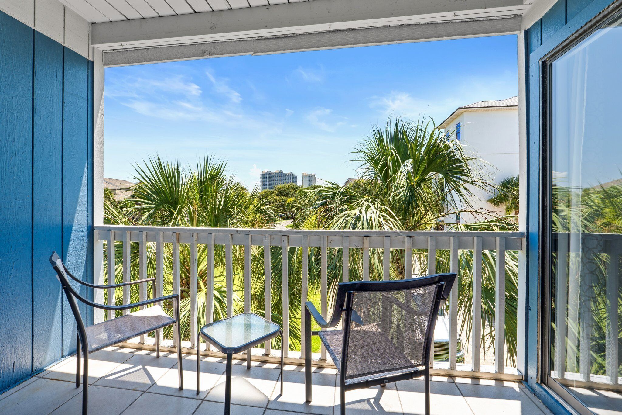 A balcony with two chairs and a small table, overlooking a cityscape with palm trees and buildings.