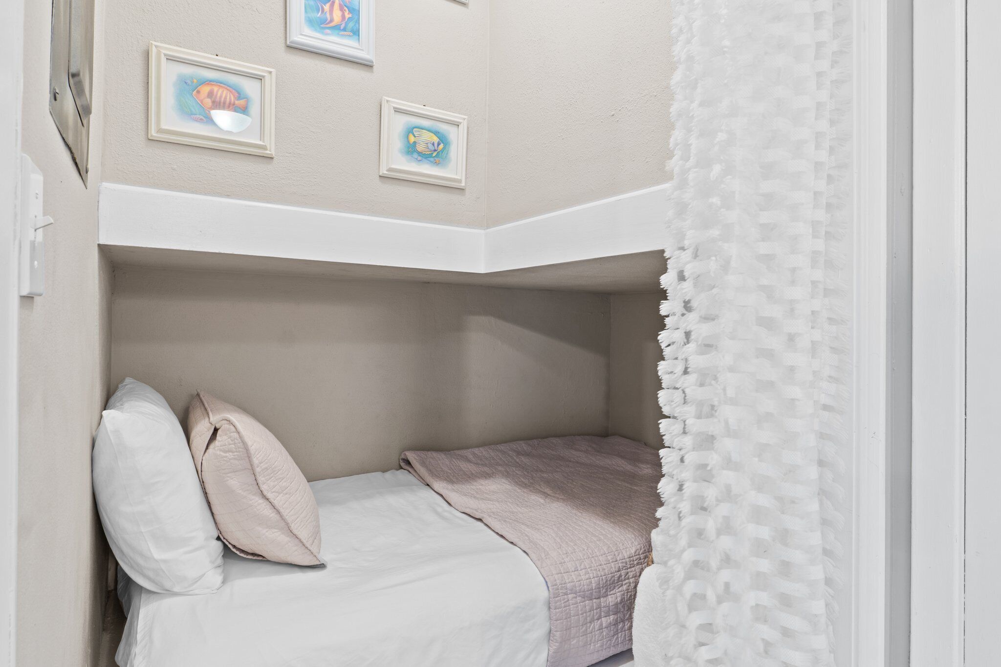 Una cama litera con cabecera blanca, manta rosa y dos almohadas. Encima de la cama hay cuadros en la pared.