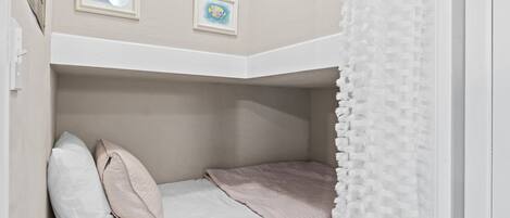Una cama litera con cabecera blanca, manta rosa y dos almohadas. Encima de la cama hay cuadros en la pared.