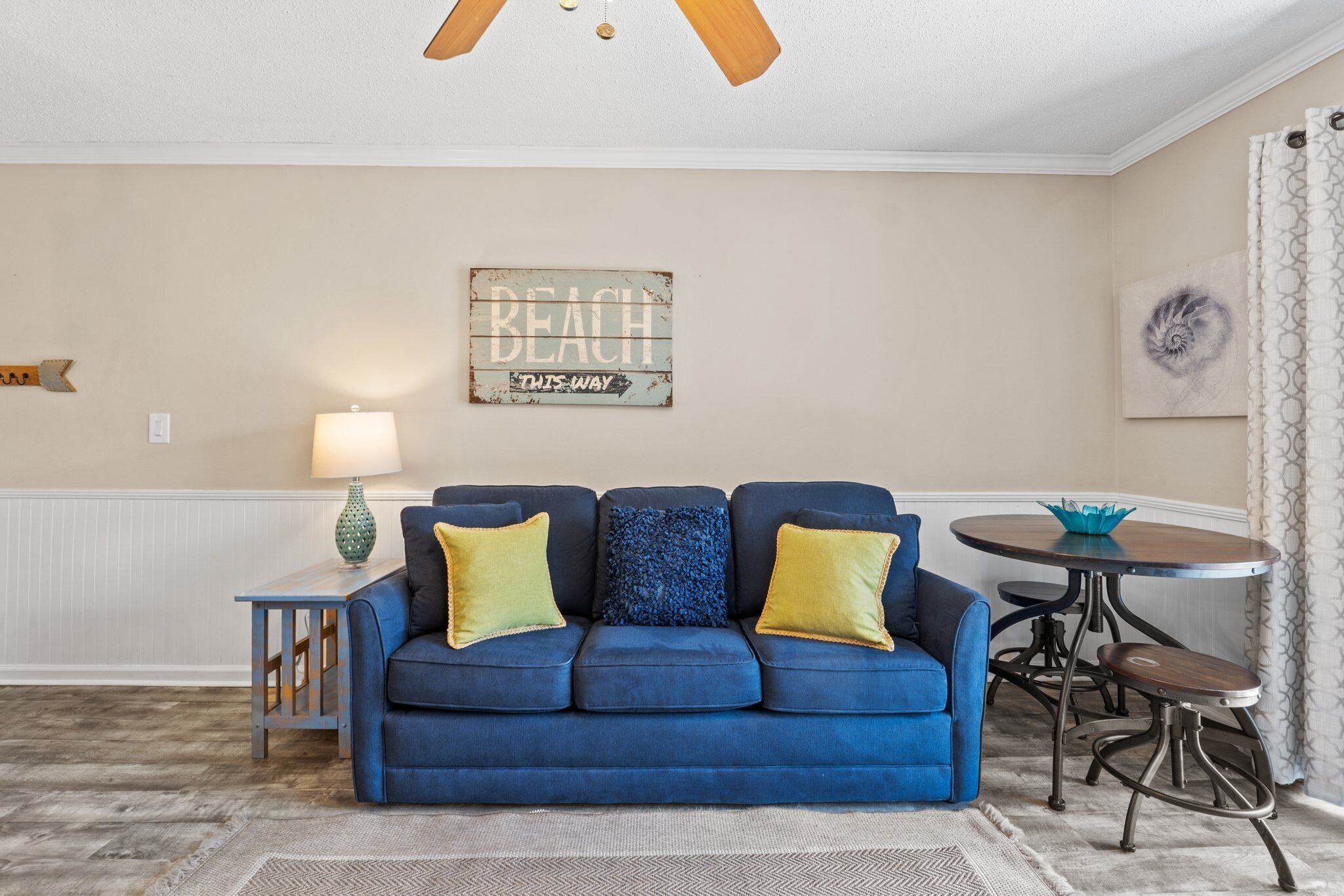 Un salon avec un canapé bleu, une petite table, une lampe et un panneau mural sur lequel on peut lire : « BEACH OUTS WAY ».
