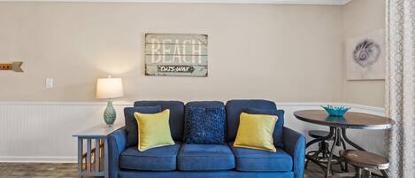 A living room with a blue sofa, a small table, a lamp, and a wall sign that reads "BEACH OUTS WAY".