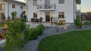 Une maison à deux étages avec un balcon, une terrasse dotée d’une table et de chaises, et un jardin bien entretenu.