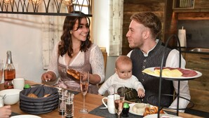 Une famille est assise autour d’une table en bois, devant différents plats, des verres et un panier à pain.