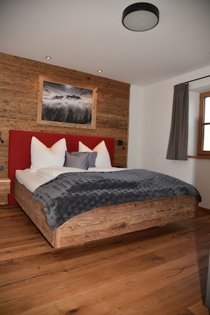 Una cama con estructura de madera, cabecera roja, almohadas blancas y grises, y manta gris.