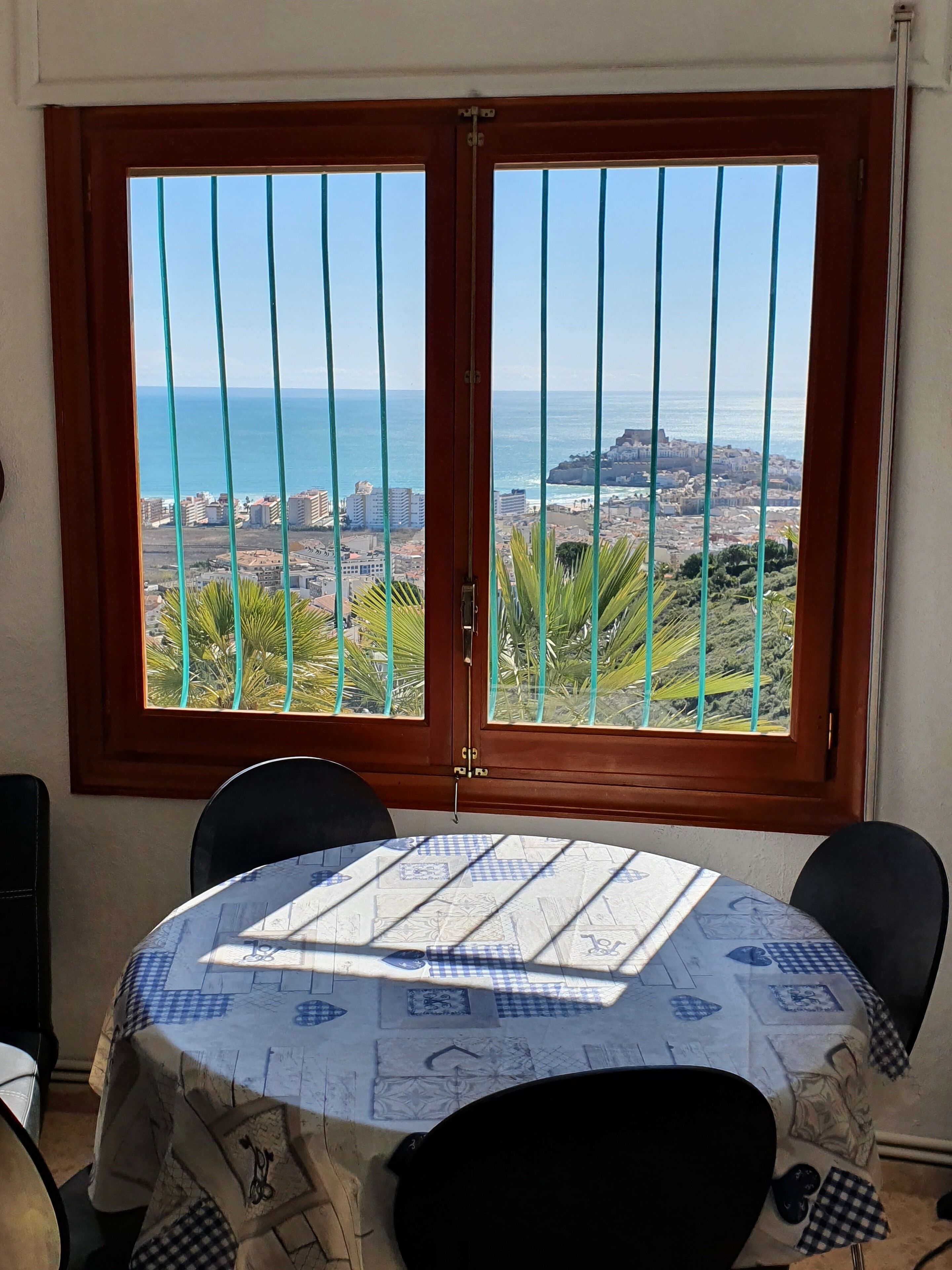 Un coin repas avec une table dressée pour deux personnes, une fenêtre donnant sur la mer et un paysage rocheux, et une porte équipée d’une barre de sécurité en métal.