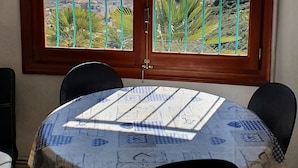 Een eethoek met een tafel gedekt voor twee personen, een raam met uitzicht op de zee en een rotsachtig landschap, en een deur met een metalen veiligheidsbalk.