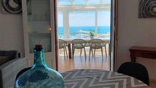 Een kamer met een groot raam met uitzicht op zee, een eethoek met stoelen en een tafel, en een decoratieve glazen fles op een geruit tapijt.