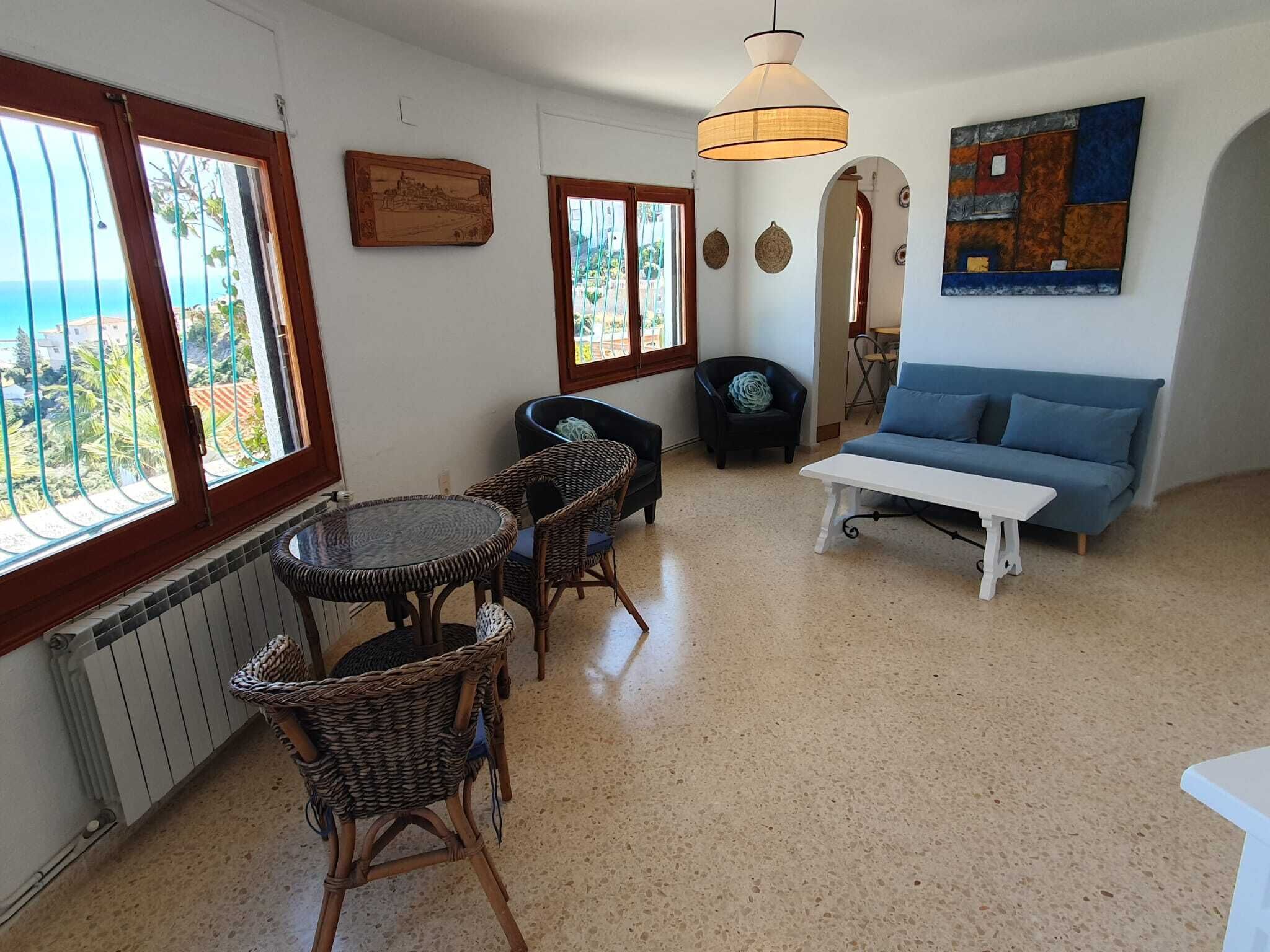 En stue med havudsigt, en blå sofa, fletstole, et hvidt bord og en hængende lampe.