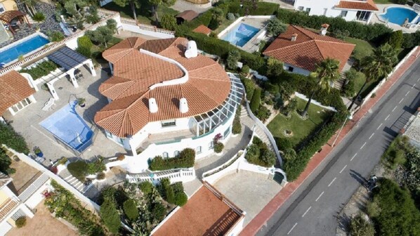 An aerial view of a residential area with a curved-roofed house, a swimming pool, and a road.
