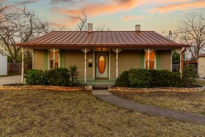A single-story house with a gabled roof, a front porch with a green door, and a small front yard with a pathway leading to the entrance.