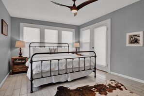 A bedroom with a metal bed frame, white bedding, a cowhide rug, a ceiling fan, and a nightstand with a lamp.