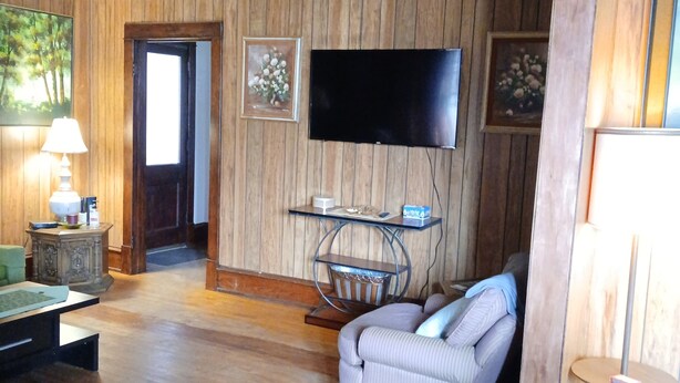 Ein Zimmer mit Holzvertäfelung, an der Wand ein Flachbildfernseher, ein kleiner Tisch mit Lampe, ein gepolsterter Sessel und ein Sofa.