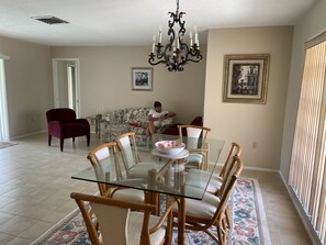 Un coin repas avec une table à plateau en verre, des chaises en osier, un lustre et un canapé en arrière-plan.