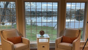 A room with two beige armchairs, a small white side table with a decorative item, and a view of a lake through large windows. - Tranquil  Beautiful Waterfront  Near Historic St. Michaels & Royal Oak (easton)