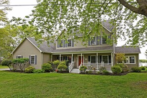 A two-story house with a front porch, a red door, and a small front yard with various shrubs and a tree.