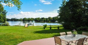 A lakeside patio with a table and chairs, a wooden deck, and a view of the lake and trees.