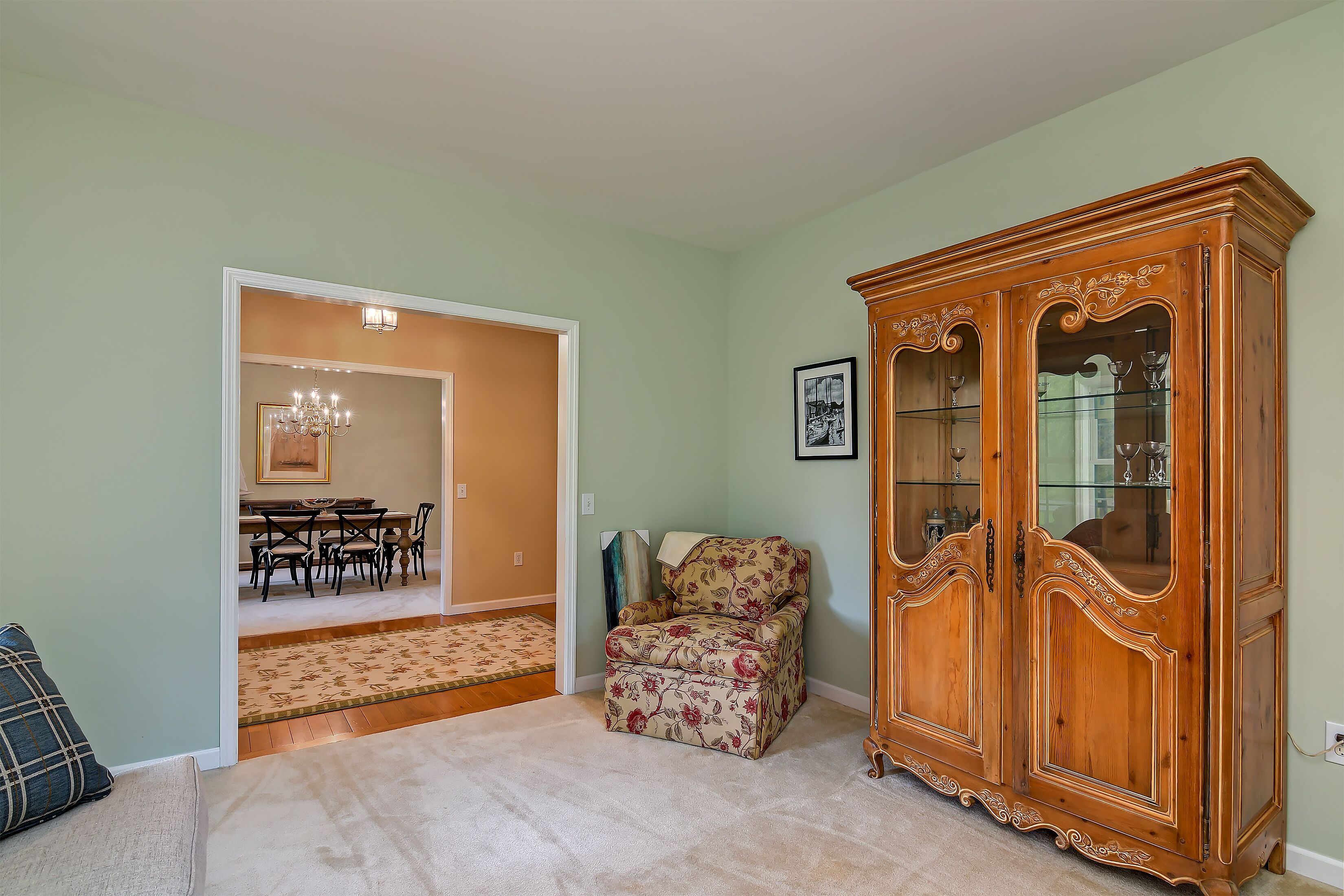 A room with a wooden cabinet, a floral armchair, a dining area with a chandelier, and a carpeted floor.