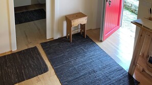 A room with a wooden ceiling, a white door, a red door, a small wooden stool, a black rug, and a wooden shelf with a vase and a basket. - Unique Experience in the Heart of Nature (Chãos)