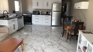 A compact kitchen with white cabinets, a refrigerator, a stove, and a dining area with a wooden table and chairs. - Aruba 2-bedroom duplex for rent. 4 minutes to Eagle Beach and close to shopping. (Noord)