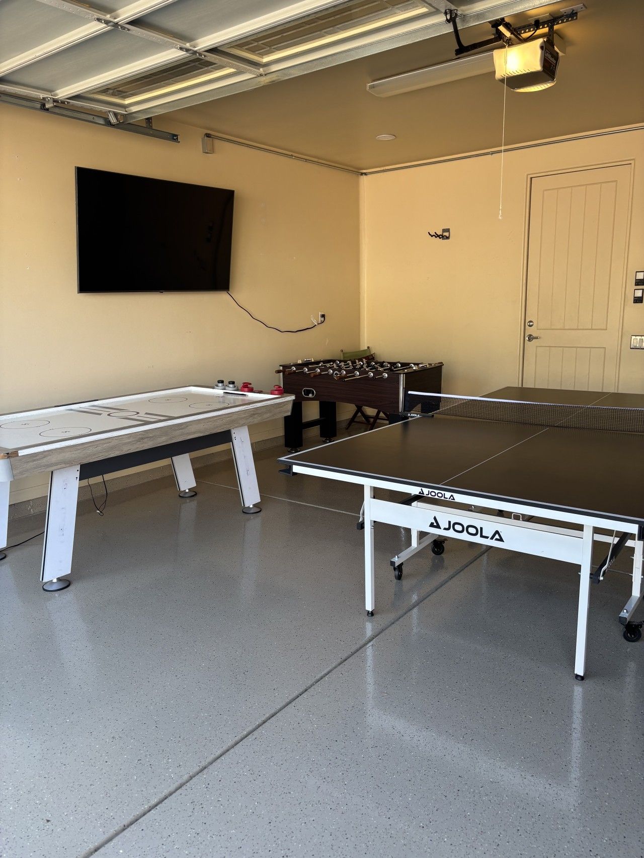 A ping pong table with a net, a foosball table, and a television mounted on the wall.