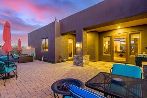 An outdoor patio area with seating and a fire pit, a modern building with a stone accent wall, and a sunset sky.