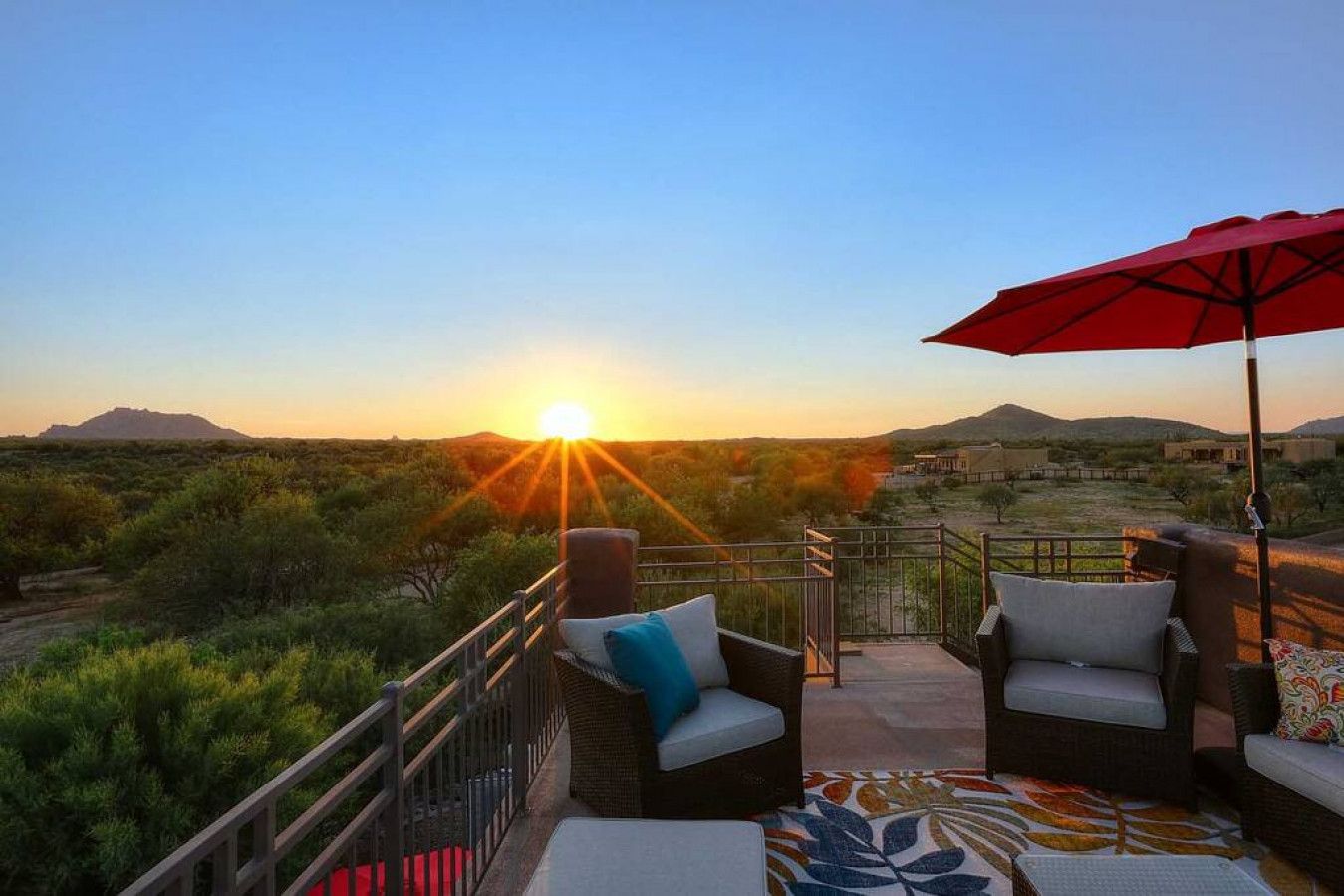 A balcony with wicker furniture and a red umbrella, overlooking a desert landscape with a sunset.