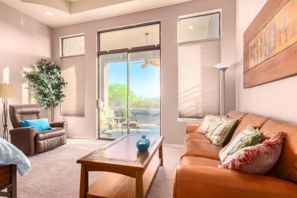 A modern living room with a brown leather sofa, a wooden coffee table, and a brown leather chair with a blue pillow. There is a large window with blinds, a potted plant, and a painting on the wall. - Desert Tranquility 4 Bedroom with Heated Pool (Scottsdale)