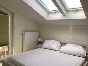 A bedroom with a skylight, a bed, a nightstand, a lamp, and a window with blinds. - 2 balconies directly by the sea, living room, fireplace, 1 bedroom, kitchen, modern bathroom with shower/WC, washing machine. (Glowe)