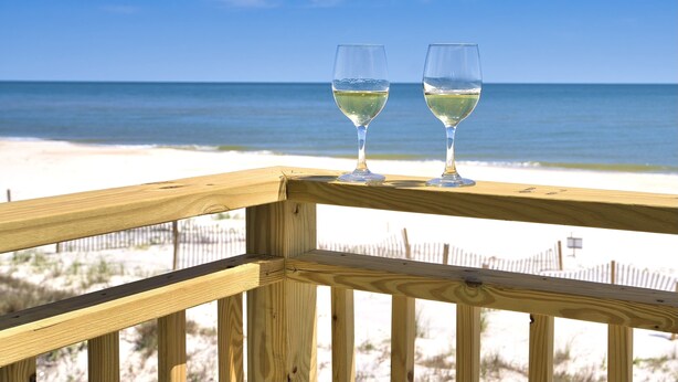 A wooden deck with two glasses of white wine on a beach with a fence in the background.