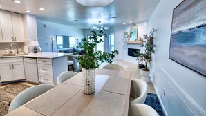 A modern kitchen with white cabinets, a dining area with a wooden table and chairs, and a living room with a fireplace and a large painting.
